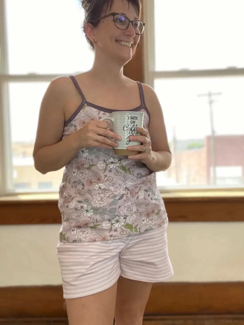 A woman wears a mauve floral tank top and striped shorts, holding a coffee mug.