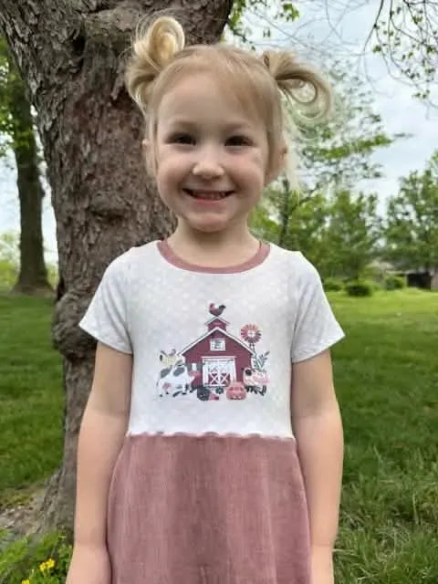 A young girl smiles, wearing a dress with a barn and farm animals on the bodice.