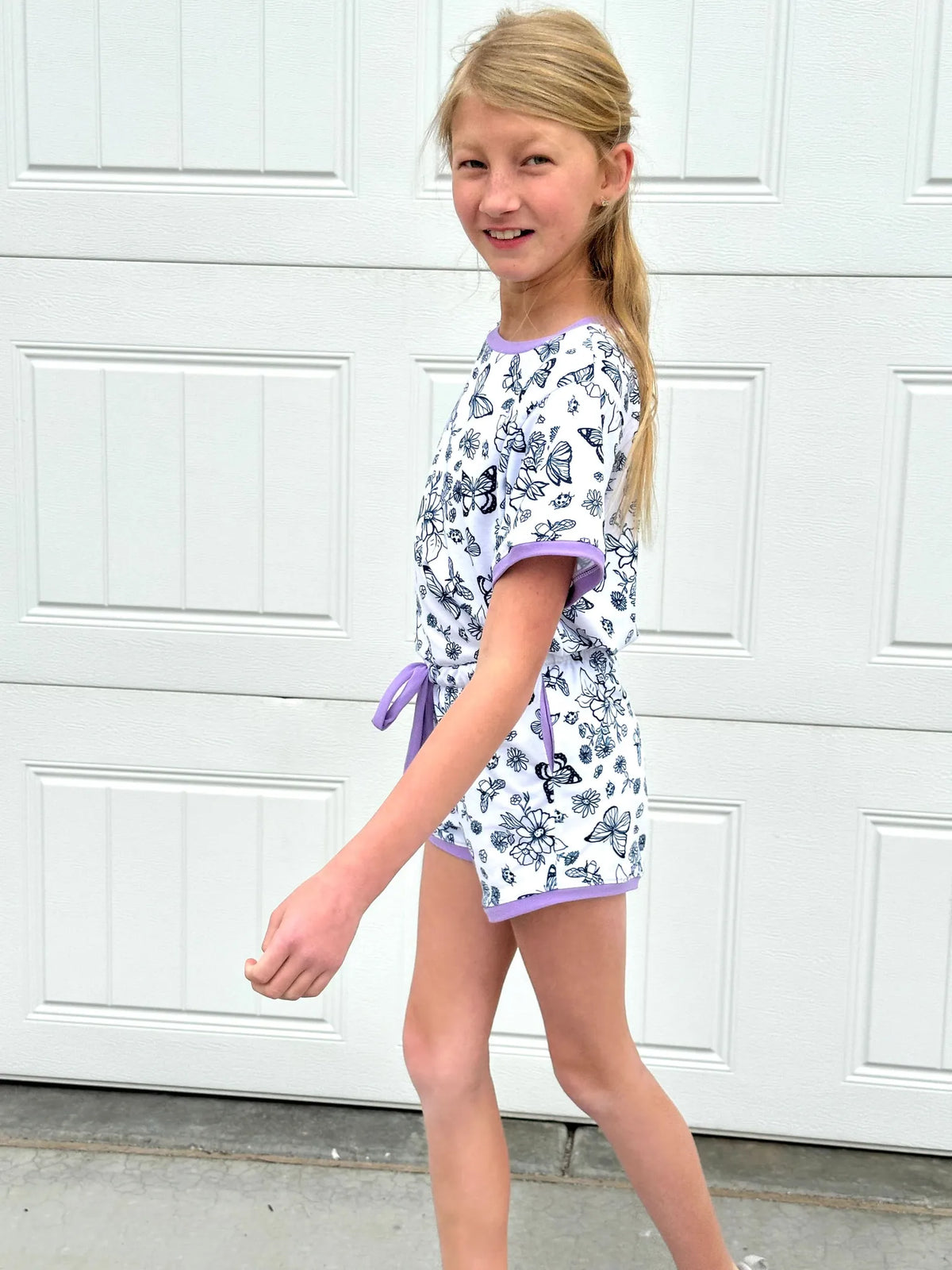 A young girl wears a white romper with black butterfly and flower outlines and purple trim.