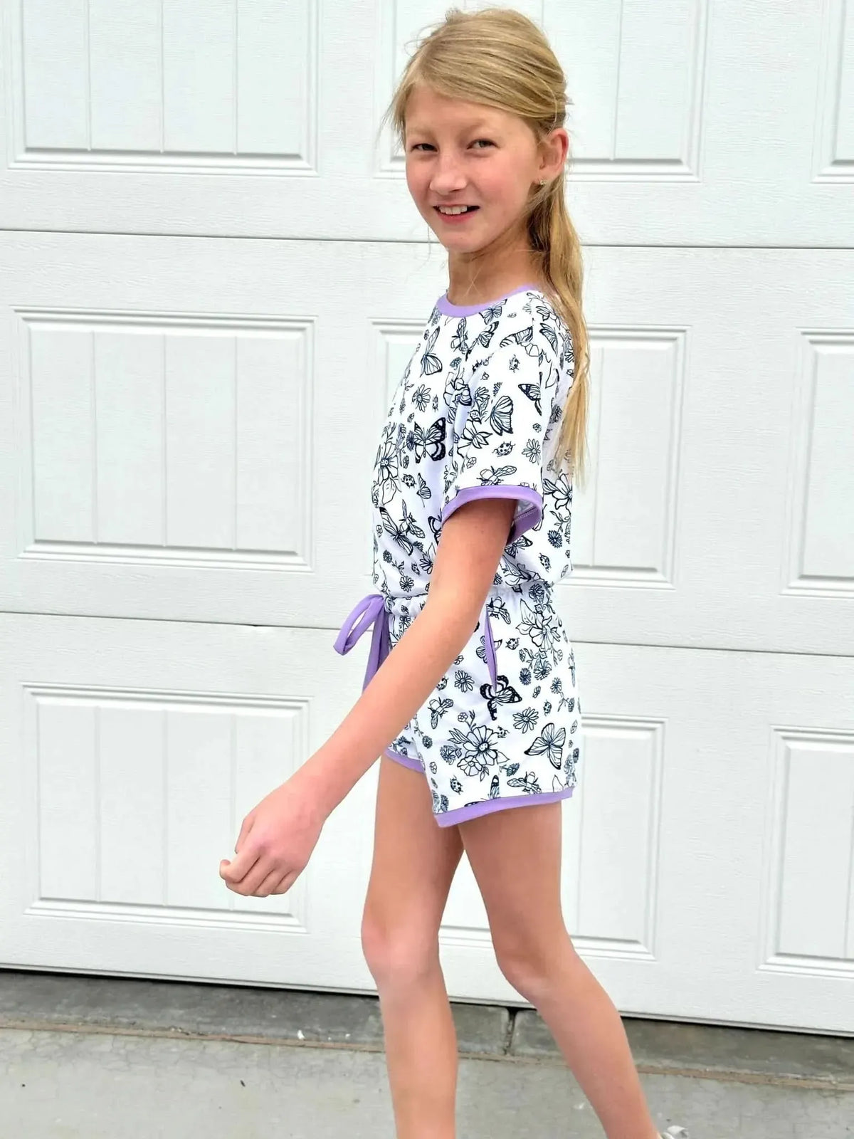 A young girl wears a white romper with black butterfly and flower outlines and purple trim.