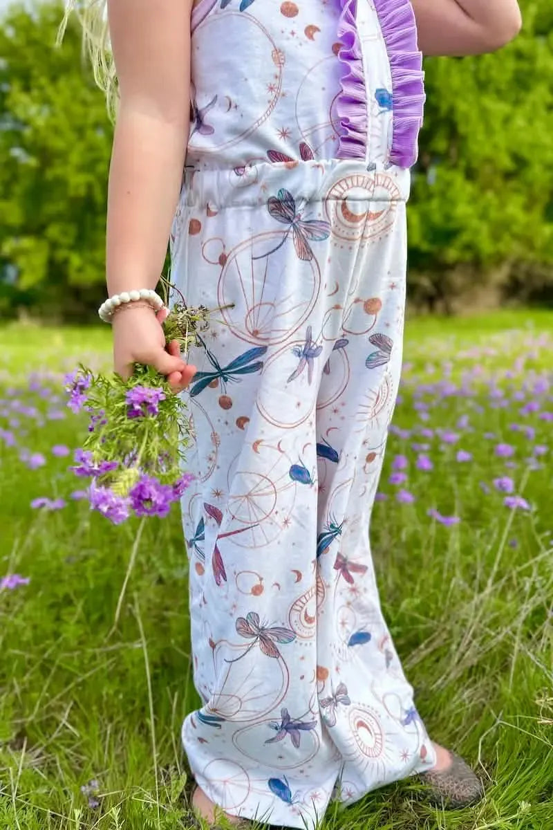 A child in a white jumpsuit with celestial dragonfly print holds purple flowers in a field.