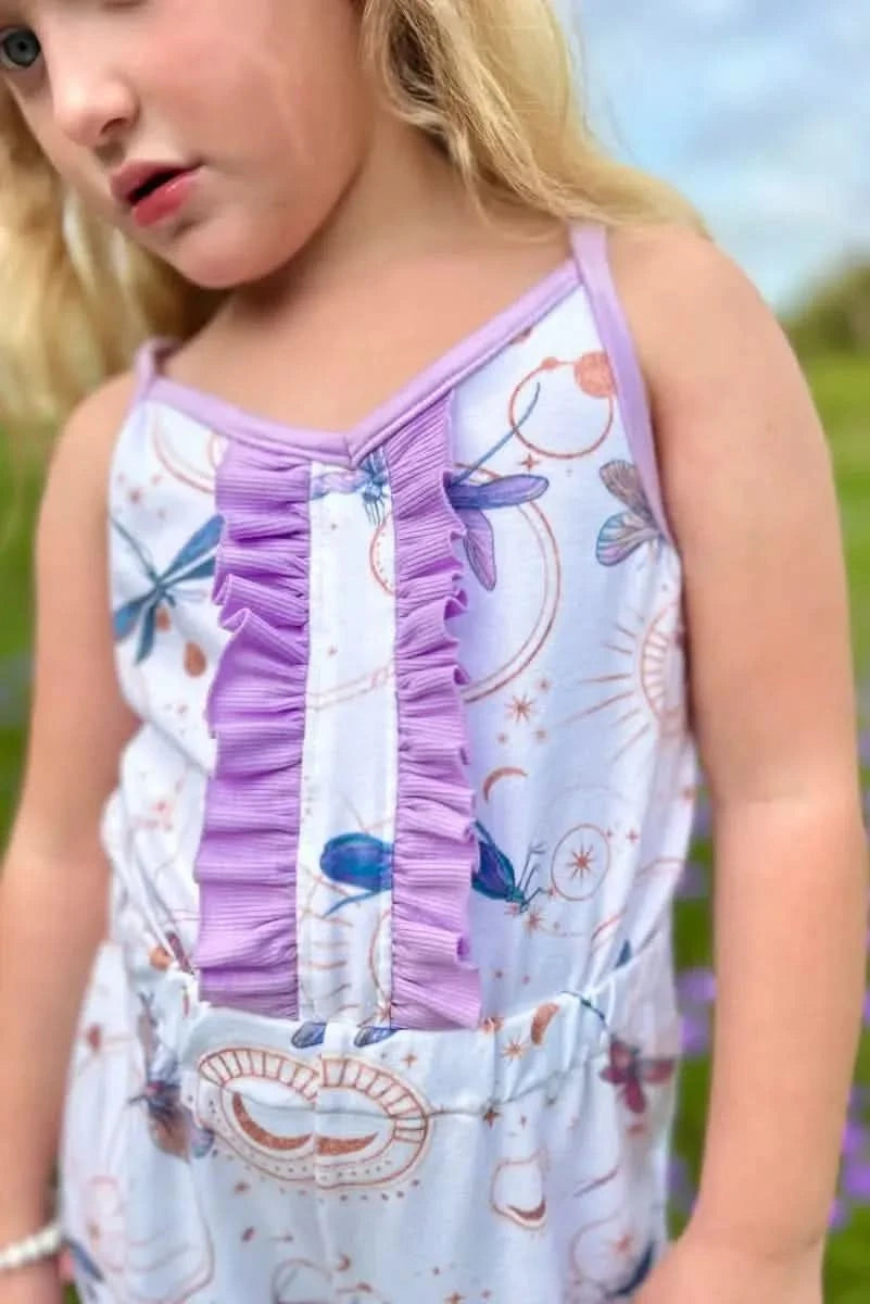 A young girl wears a white romper with celestial dragonfly print and purple ruffles.