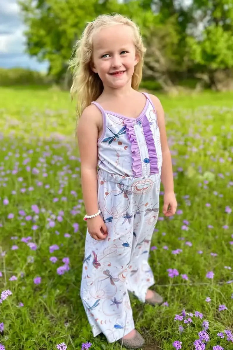A young girl wears a white jumpsuit with purple ruffles and dragonfly print.