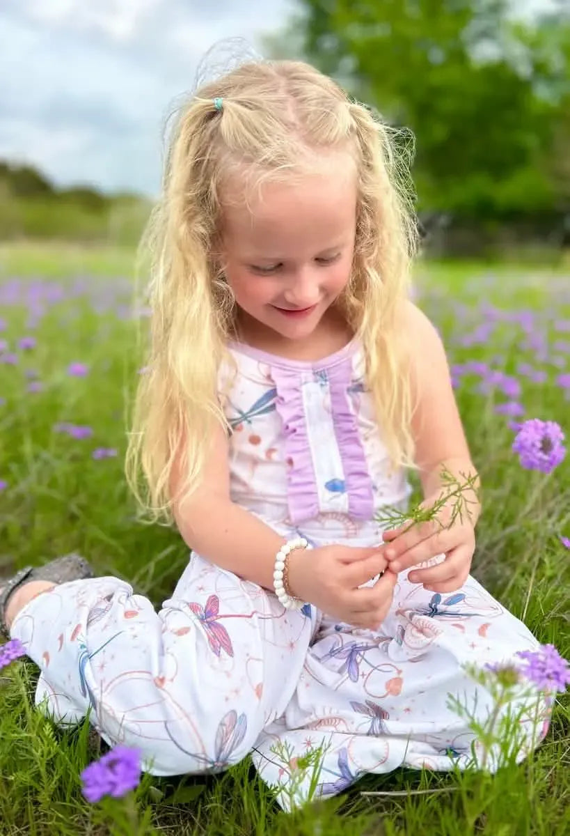 A young girl in a white outfit with celestial dragonfly print sits in a field of purple flowers.