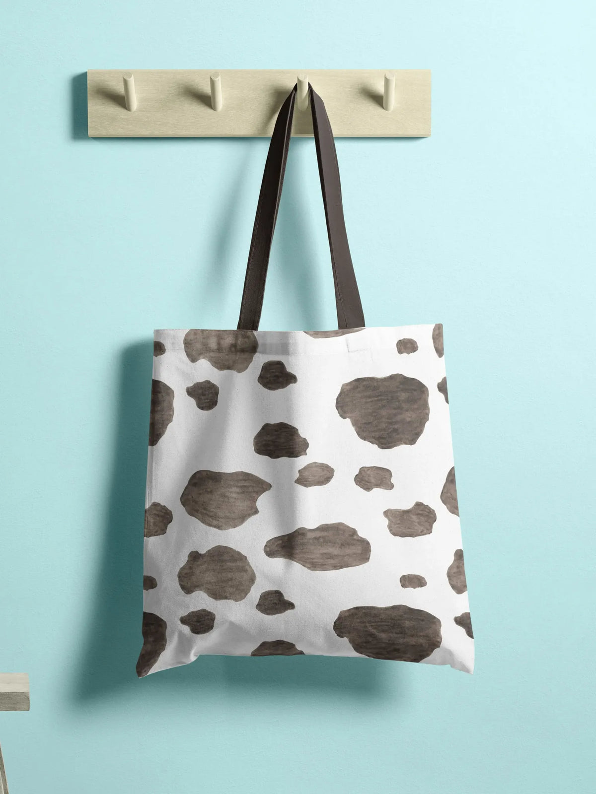 A white tote bag with brown cowhide spots hangs on a wooden rack.