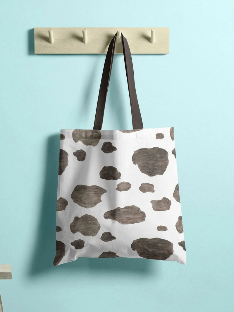 A white tote bag with brown cowhide spots hangs on a wooden rack.