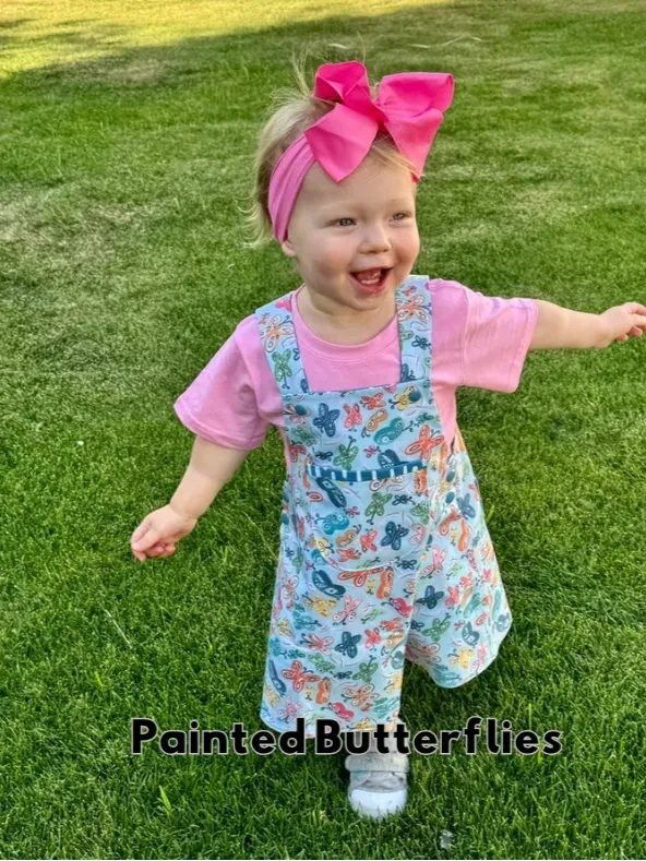 A toddler in a pink shirt and butterfly-print overalls with a large pink bow.