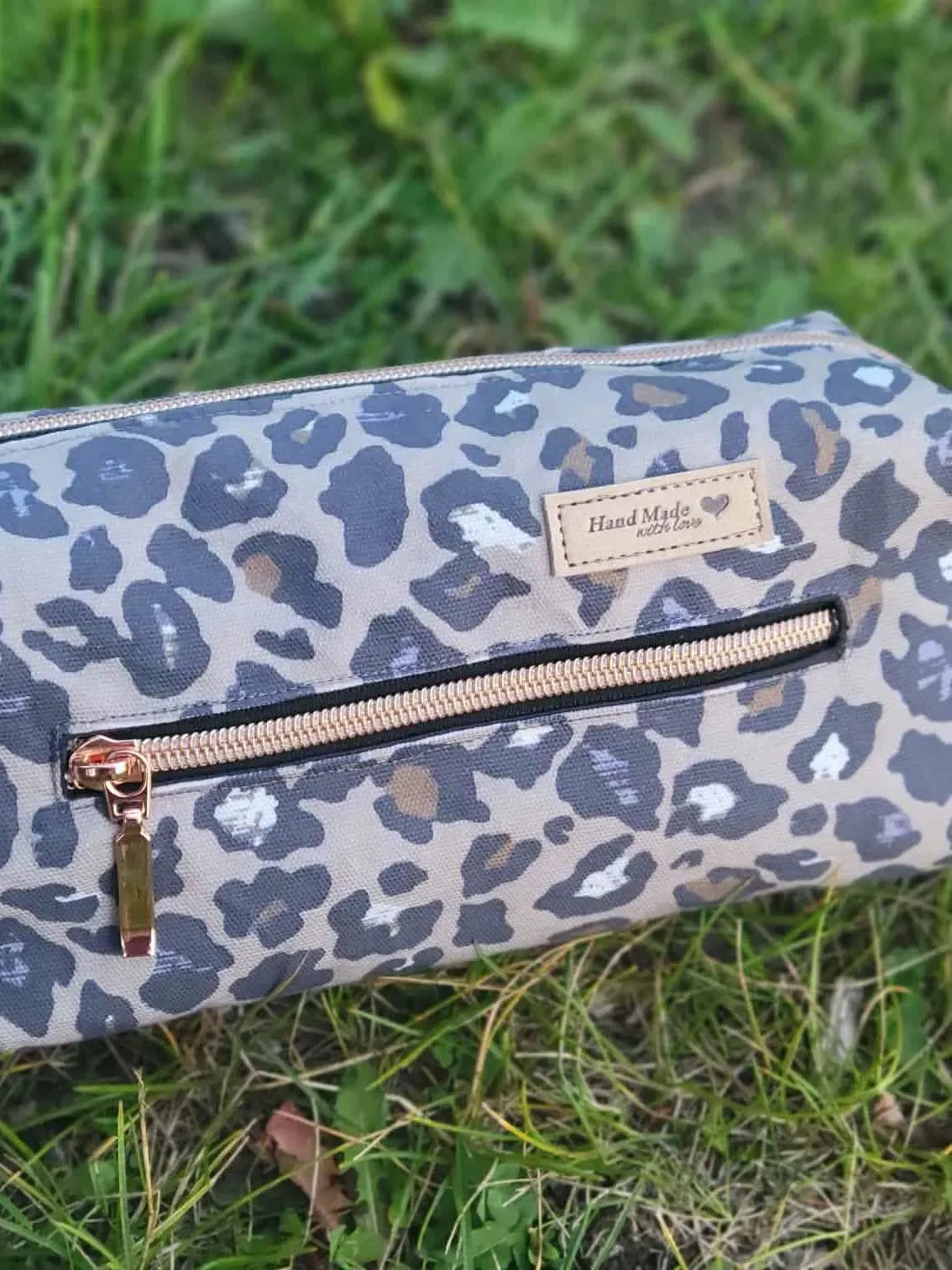 An Autumn Leopard print pouch with a "Hand Made with love" tag lies on grass.