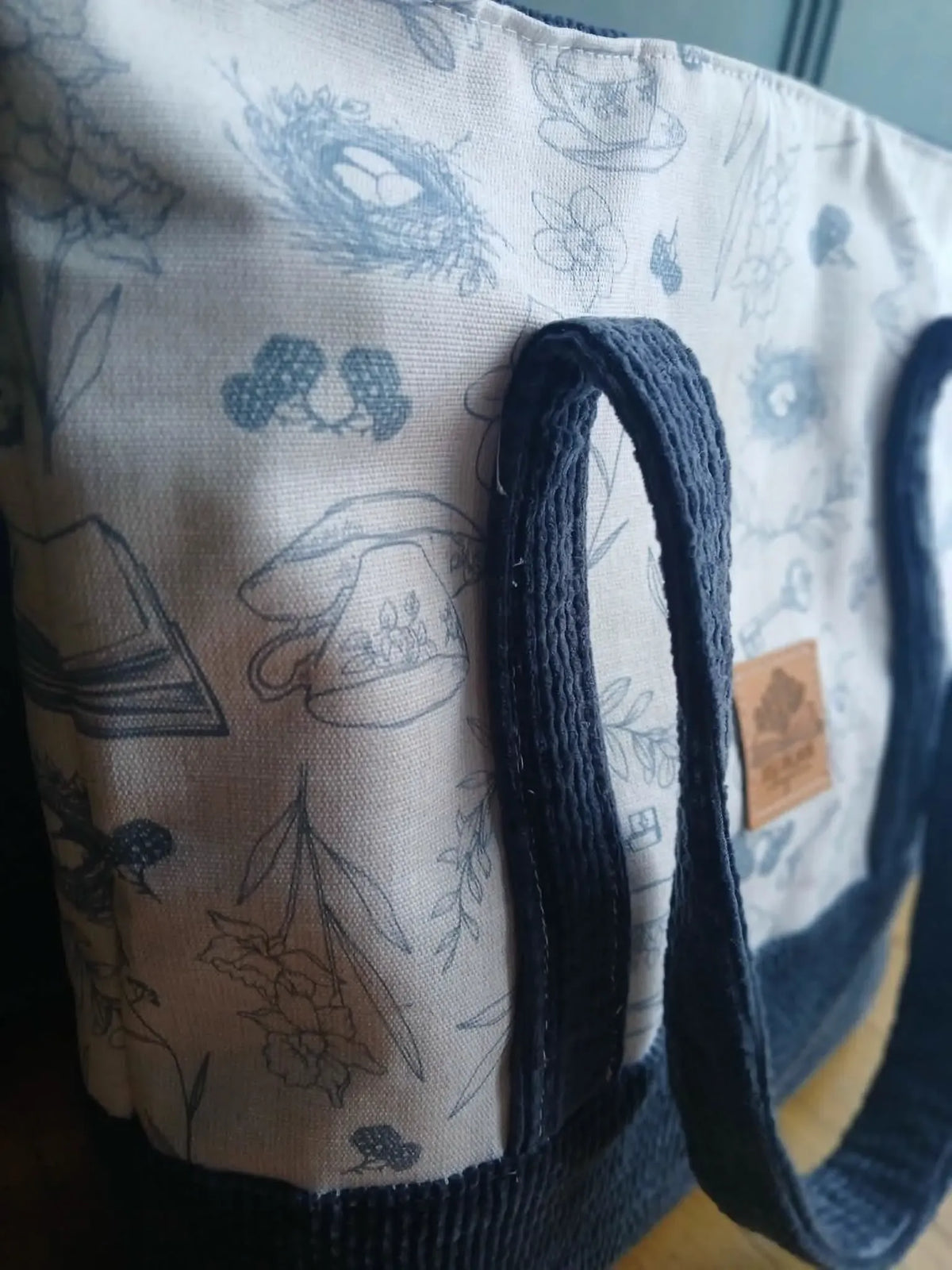 A canvas tote bag with blue illustrations of nature and tea items, and dark blue handles.