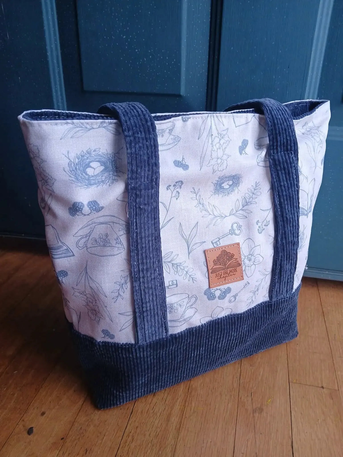 A white tote bag with blue toile patterns and a brown leather tag.