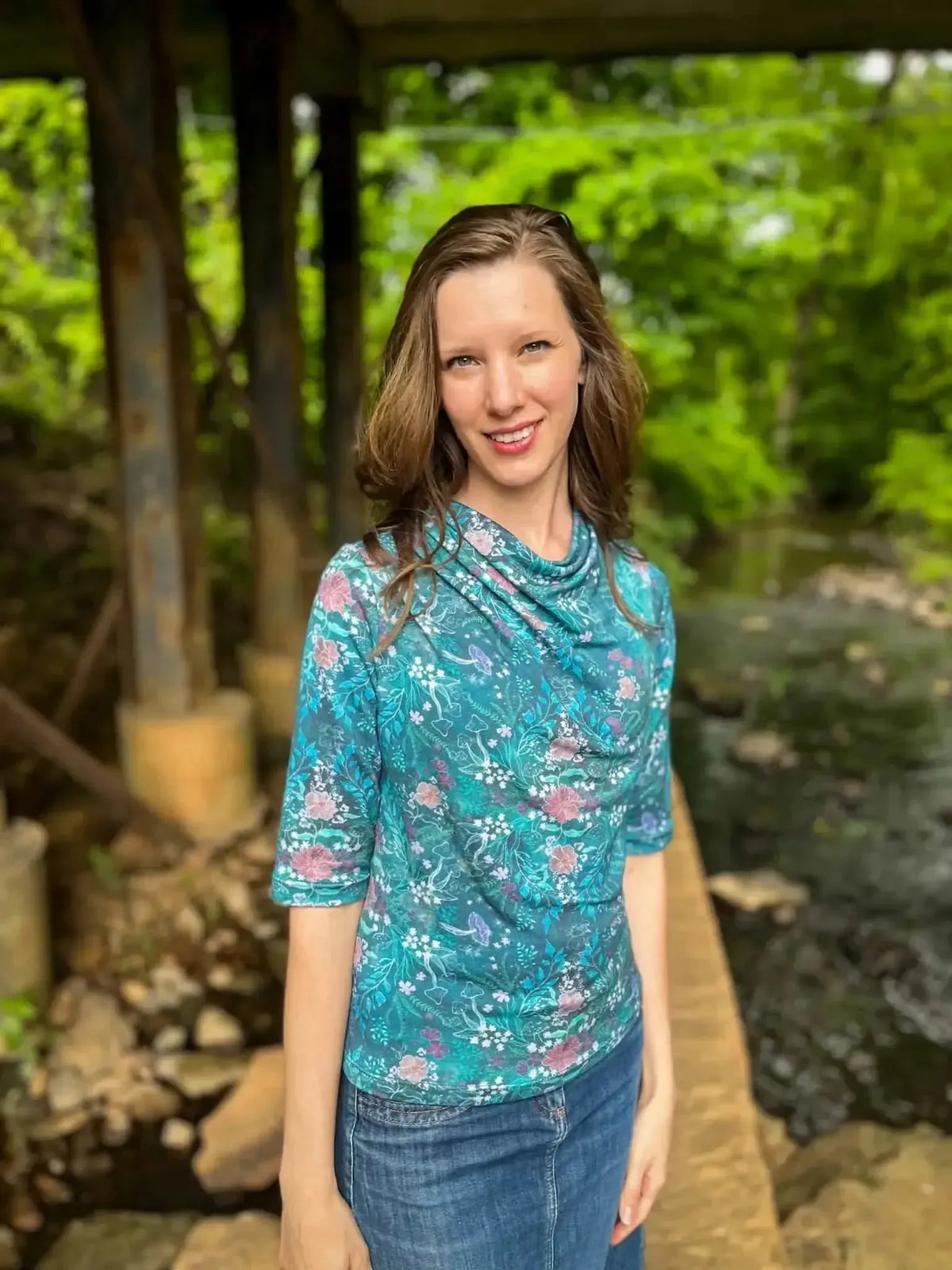 A woman wears a teal woodland print top and jeans by a stream.