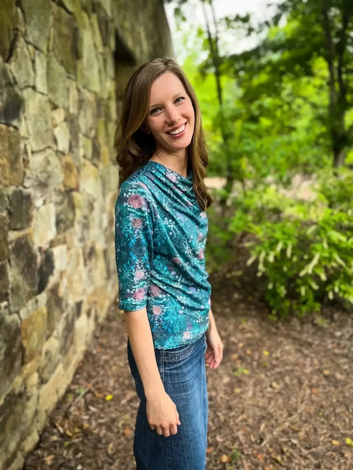 A woman smiles, wearing a teal woodland print top and denim skirt.