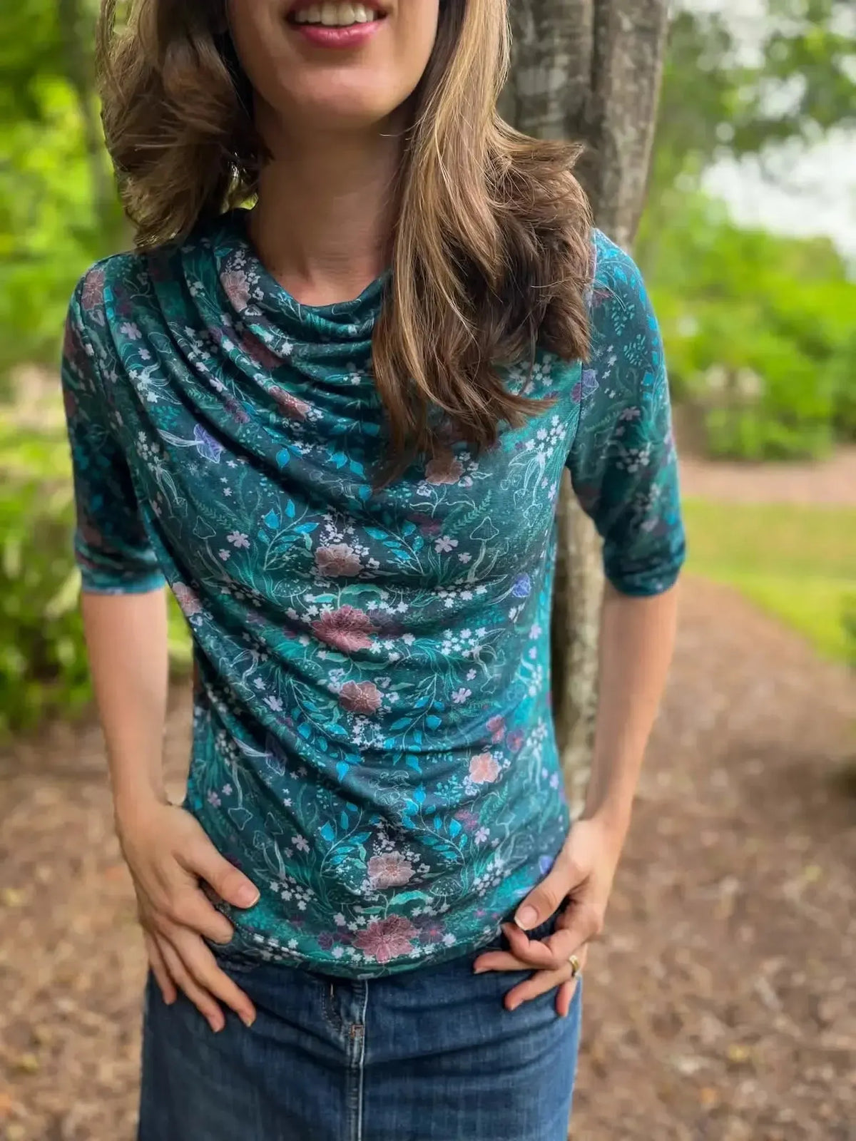A woman wears a teal woodland print top with a cowl neck and denim skirt.