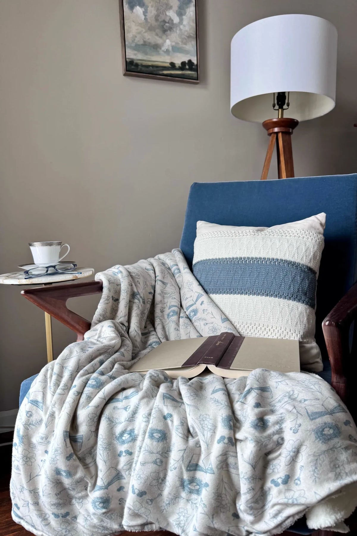 A cozy blue armchair with a patterned blanket, pillow, and open book.