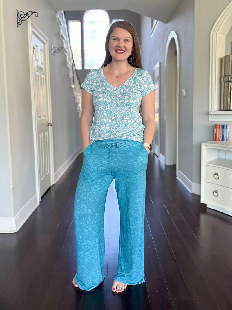 A woman smiles, wearing a floral v-neck top and dark teal heather wide-leg pants.