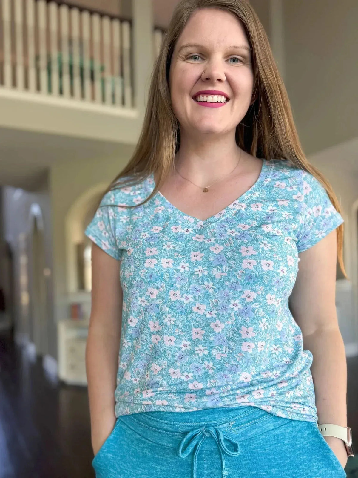 A woman smiles, wearing a light blue floral V-neck shirt and blue shorts.