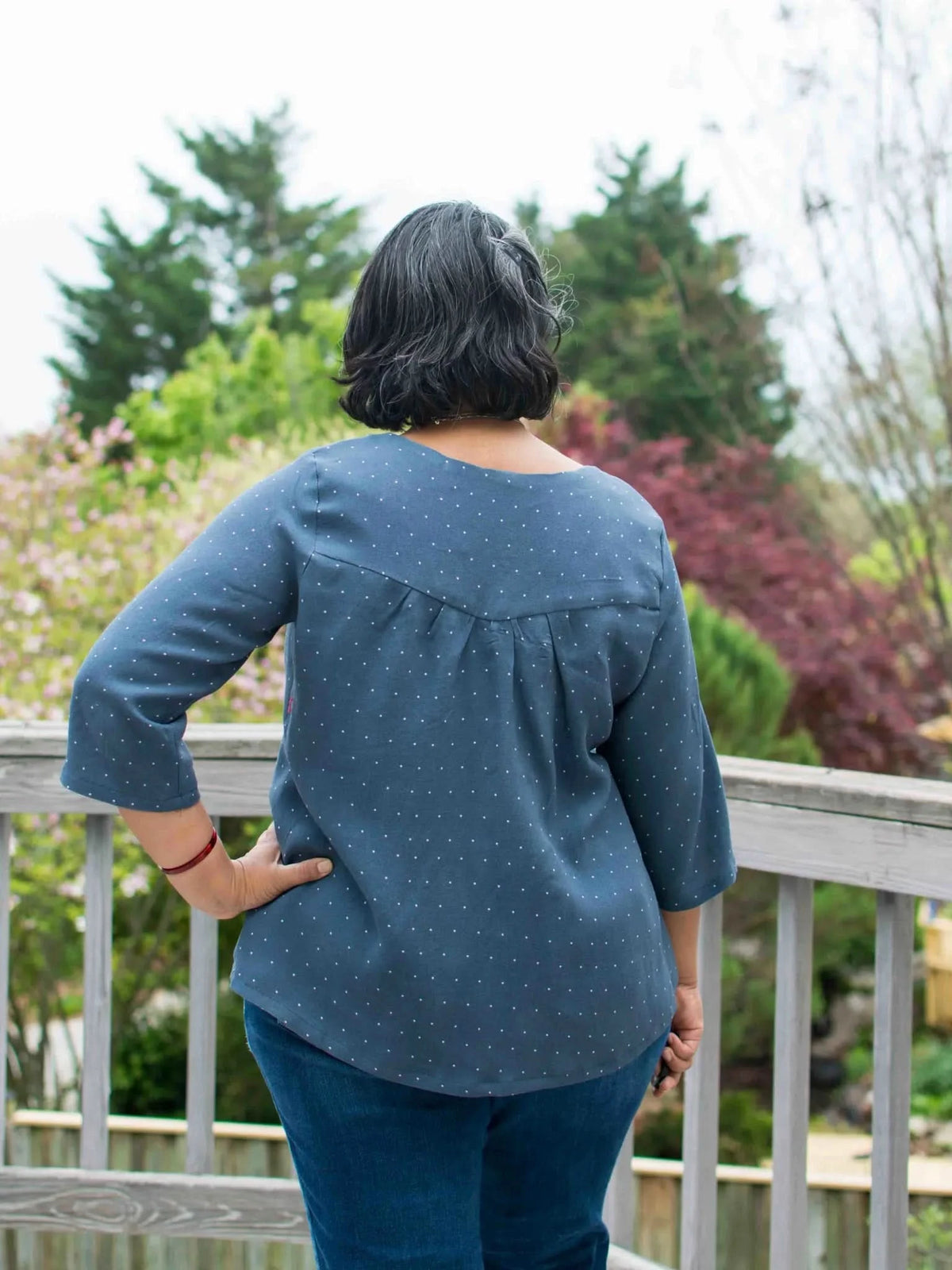 A person from behind wears a navy polka dot top and jeans on a wooden deck.