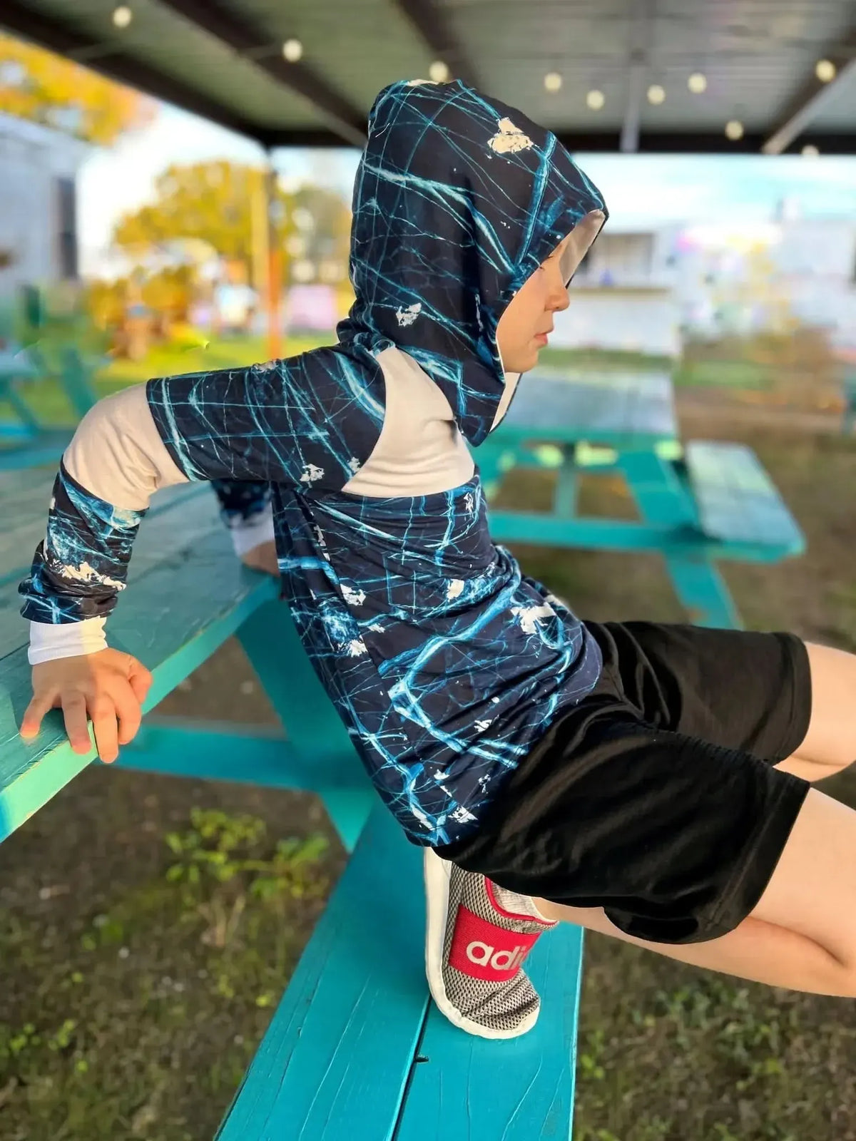 Child in a navy cracked ice hoodie and black shorts sits on a blue picnic table.