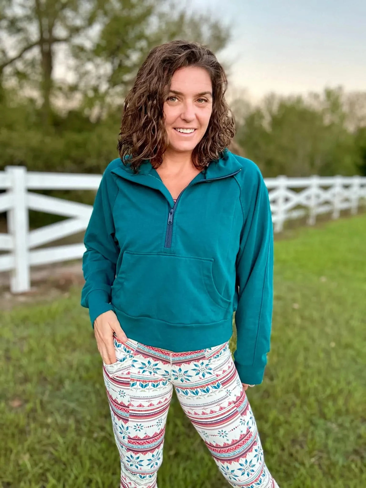 A woman in a teal hoodie and patterned leggings stands outdoors.